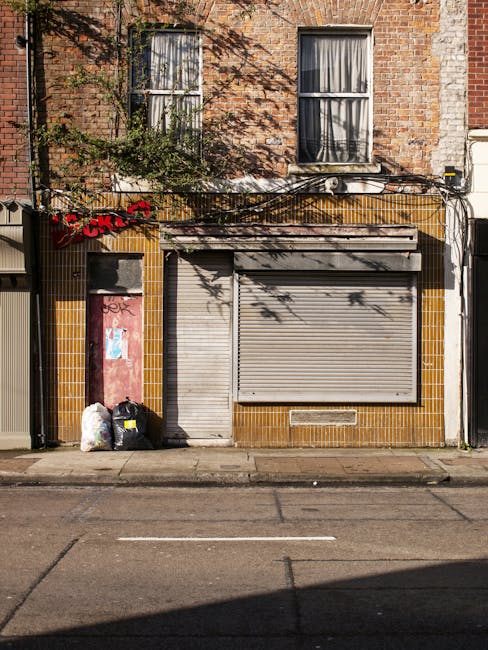 Two large black plastic rubbish bags, slightly crumpled and tied at the top, are placed on the pavement near a black metal fence with pointed vertical bars. The bags appear to contain mixed waste mate